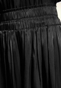 Jupe plissée noire en tissu brillant avec une taille froncée, présentant des plis verticaux et une texture lisse.