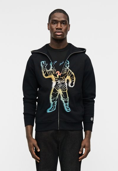 Άνδρας με μαύρο φούτερ με φερμουάρ και πολύχρωμο γραφικό αστροναύτη, φορεμένο με μαύρο παντελόνι, στέκεται μπροστά σε απλό ανοιχτό γκρι φόντο.