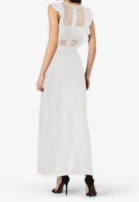Biała sukienka maxi z falbaniastymi rękawami, przezroczystym tyłem z siateczki, plisowanym dołem i dopasowaną talią. Noszona z czarnymi butami na wysokim obcasie.