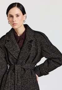 Cappotto trench in lana testurizzata bianco e nero con ampio colletto, vita con cintura e spalline imbottite. Presenta grandi bottoni su una camicia scura sottostante.