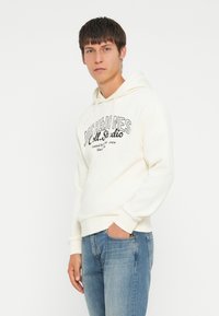 Cremefarvet hoodie med sort grafisk skrifttype; blødt materiale, kengurulomme, snoretræk i hætten, ribbede manchetter, båret med blå jeans.