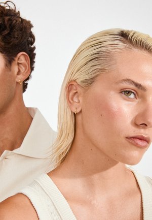 Zlaté kruhové náušnice zdobia uši modelky s krátkymi blond vlasmi, ktorá predvádza hladkú štruktúru pleti a neutrálny make-up. Pozadie je jednoduché.