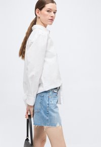 Νεαρή γυναίκα με μακριά καστανά μαλλιά σε αλογοουρά, ντυμένη με λευκό πουκάμισο με κουμπιά και μίνι φούστα από σκισμένο τζιν, κρατώντας μαύρη τσάντα χειρός.