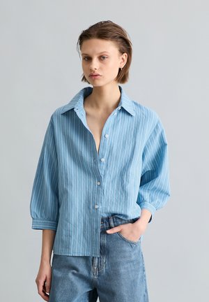 Młoda osoba z krótkimi włosami, ubrana w luźną, jasnoniebieską, w paski koszulę zapinaną na guziki oraz niebieskie dżinsy, z jedną ręką w kieszeni na jednolitym tle.