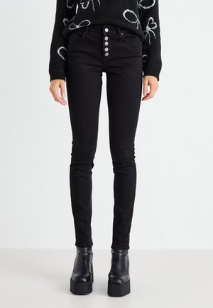 Personne portant un jean skinny noir taille haute avec une braguette à cinq boutons et des bottines noires à plateformes, debout devant un fond blanc uni.