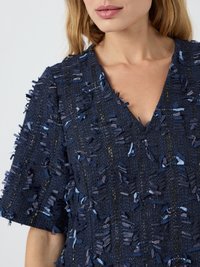 Artimųjų planas, kurioje matoma moteris, vilkinti tamsiai mėlyną tekstūruotą megztinį su trumpomis rankovėmis ir V formos iškirpte, puošniai išmargintą atsitiktiniais audinio pakraščiais.