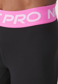 Black athletic leggings featuring a wide pink waistband with "NIKE PRO" printed in white text. Smooth texture, designed for comfort and flexibility.