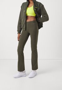 Giacca in pile verde oliva con zip, leggings aderenti verde oliva abbinati e scarpe da ginnastica in mesh bianche. Sotto si vede un reggiseno sportivo giallo brillante.