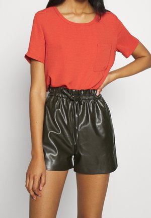Femme portant une chemise rouge vif à manches courtes avec une poche avant et un short en cuir noir taille haute à cordon de serrage.