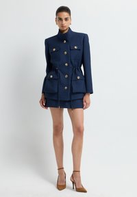 Giacca texturizzata blu navy con bottoni dorati, vita regolabile con cordoncino e due tasche frontali, abbinata a una mini gonna coordinata e tacchi marroni.