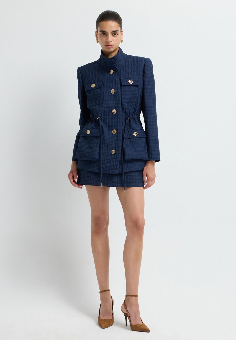 Giacca texturizzata blu navy con bottoni dorati, vita regolabile con cordoncino e due tasche frontali, abbinata a una mini gonna coordinata e tacchi marroni.