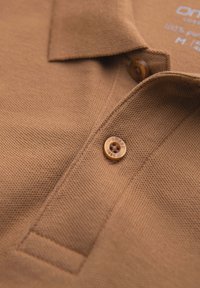Nærbillede af kraven på en beige polo-shirt med tekstureret stof og mærket brun knap, som lukker slidsen.
