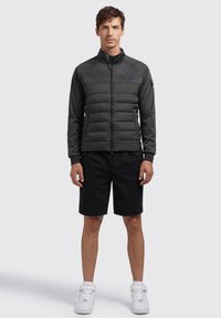 Men's jacket featuring a padded gray body with a smooth, dark gray upper section and a high collar. Paired with black shorts and white shoes.