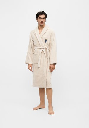 Homme portant un peignoir beige jusqu'aux genoux avec ceinture et petit ours brodé sur la poche poitrine, debout pieds nus sur fond blanc.
