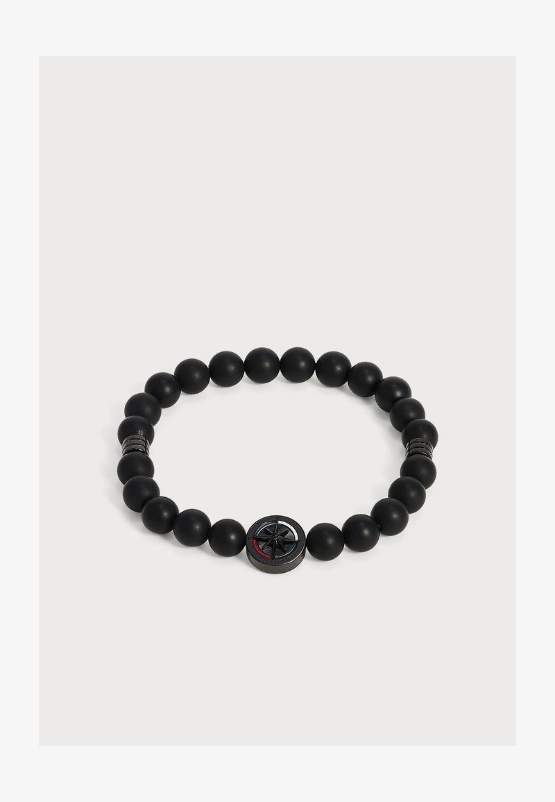 Bracelet noir à perles avec un pendentif boussole rond et deux petits pendentifs cylindriques sur un fond clair.