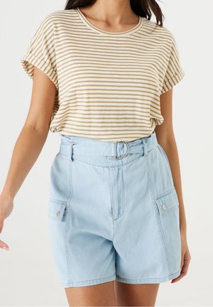 Kobieta ubrana w beżowo-białą pasiastą koszulkę z krótkim rękawem, wciętą w wysokowalne jasnoniebieskie dżinsowe szorty z paskiem i bocznymi kieszeniami.