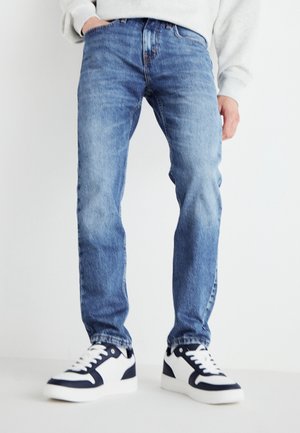 Personne portant un jean bleu en denim et des baskets blanches avec des accents noirs, debout avec une main dans la poche devant un fond clair et uni.