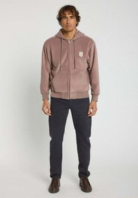 Sweatshirt à capuche zippé en polaire mauve, avec des poignets et un ourlet côtelés. Poches avant et petit logo sur la poitrine gauche. Associé à un pantalon foncé.