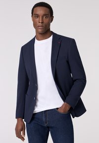 Blazer blu con texture, collo a rever e bottoni blu, abbinato a una t-shirt bianca e jeans scuri, creando un look smart-casual.