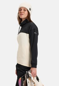 Zwarte en crème pullover met een hoge kraag, gemêleerde stof en een zak aan de voorkant. Model draagt een crème beanie en houdt met één hand uitrusting vast.
