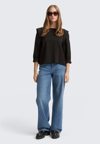 Schwarzes, strukturiertes Langarmoberteil mit Puffärmel, kombiniert mit weit geschnittenen blauen Jeans und klobigen schwarzen Schuhen. Hohe Sonnenbrille als Accessoire.