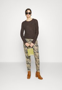 Suéter de malha castanho, calças com padrão de camuflagem, botins castanhos de salto alto e uma pequena mala verde. Os acessórios incluem óculos de sol pretos e brincos.