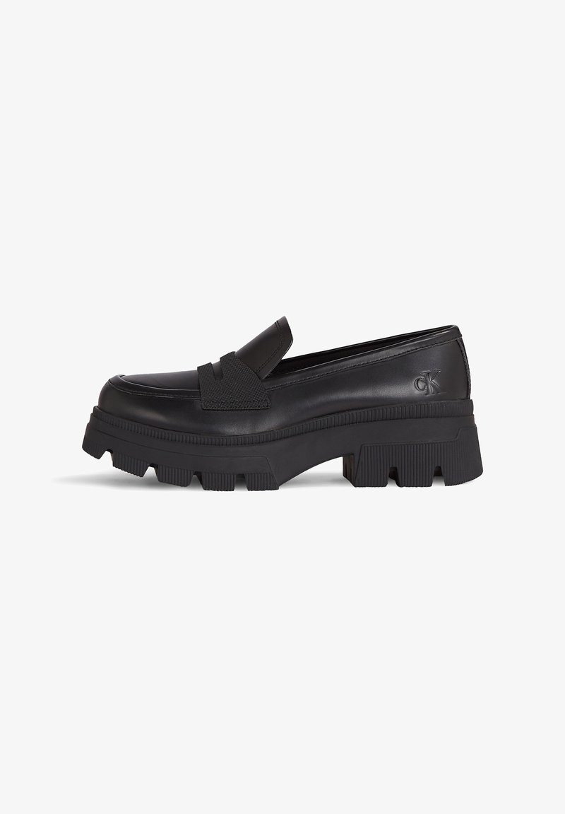 Mocassins en cuir noir avec une semelle épaisse en caoutchouc, dotés d'une bride élastique noire texturée et d'un logo embossé sur le talon.