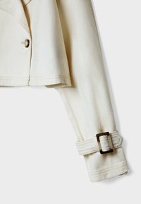 Manche de veste en denim écru avec sangle et boucle rectangulaire foncée, montrant un détail de bouton près de l’ourlet sur fond clair.