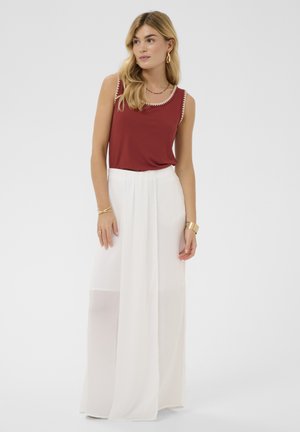Mujer con el pelo largo y rubio que lleva una camiseta sin mangas roja con ribete blanco y pantalones blancos de pierna ancha y cintura alta sobre un fondo claro y liso.