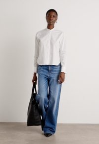 Witte katoen shirt met een hoge kraag, gecombineerd met lichtblauwe wijde jeans. Het model houdt een zwarte leren tote bag vast. Glad textuur.