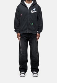 Felpa nera con zip e stampe grafiche con dettagli rossi e verdi, abbinata a jeans in denim nero dalla vestibilità ampia e scarpe da ginnastica bianche classiche.