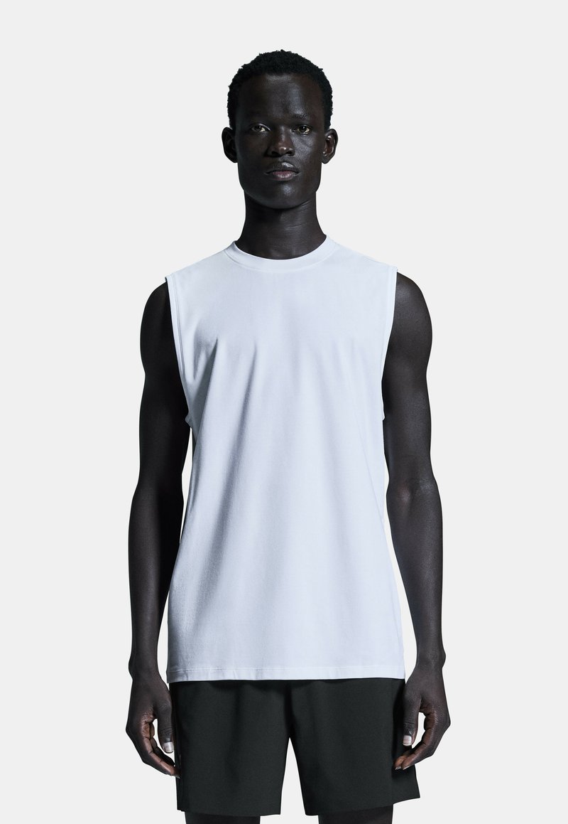 Weißes ärmelloses T-Shirt aus glattem Baumwollstoff, mit rundem Halsausschnitt und lockerer Passform, kombiniert mit dunklen Shorts.