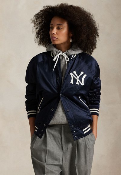Göndör hajú nő sötétkék New York Yankees szatén dzsekit visel szürke kapucnis pulóver és szürke pliszírozott nadrág fölött, kezei a zsebében, oldalra néz.
