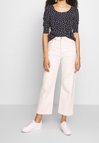 Bílé džíny s širokými nohavicemi v kombinaci s černým topem s malými bílými kruhovými vzory. Model má na sobě bílé tenisky a stojí s rukama podél těla.