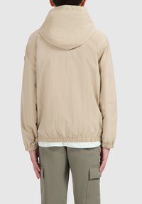 Beige lichtgewicht jacket met een capuchon, elastische manchetten en zoom, met een gestructureerd oppervlak. Draagt over een lichtgekleurde shirt en groene broek.