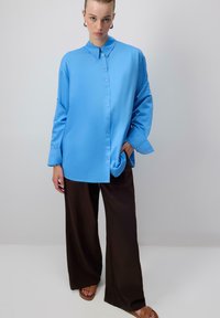 Nő simára fésült hajjal, oversized, élénk kék, gombos ingben, laza fekete nadrágban és világosbarna szandálban áll egy semleges háttér előtt.