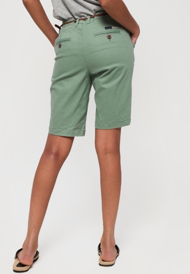 Shorts de algodón verde con corte recto, bolsillos traseros con botones y una textura suave. Llevados con una parte superior gris y sandalias.
