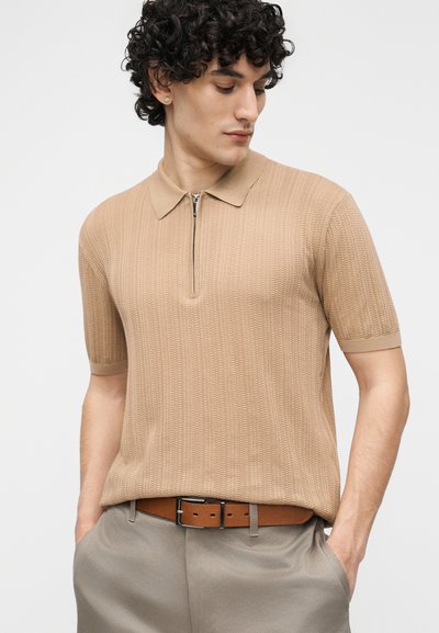 Junger Mann mit lockigem Haar, der ein beigefarbenes Polo-Shirt mit kurzem Reißverschluss, einen braunen Gürtel und graue Hosen trägt und nach unten links schaut.