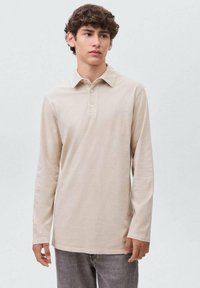 Beige polo tričko s dlouhým rukávem, knoflíkovou légou a límečkem, vyrobené z hladkého materiálu. Model má na sobě šedé kalhoty a stojí na světlém pozadí.