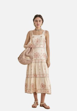 Mujer con un vestido midi sin mangas color crema con estampados florales rosas, sujetando una gran bolsa tejida, lleva sandalias y un collar y pendientes de conchas.
