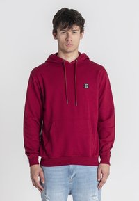 Sudadera de algodón roja con un bolsillo canguro, cordón ajustable y un pequeño parche con logo. Textura suave con puños y dobladillo acanalados.