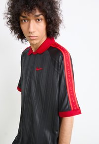 Young person with curly hair wearing a black Nike shirt with red collar and red stripes featuring Nike logos on shoulders, looking at the camera.