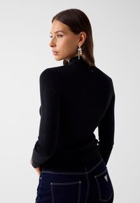 Frau mit braunen Haaren, die ein schwarzes langärmeliges Oberteil und dunkelblaue Jeans mit weißen Nähten trägt, schaut nach links vor einem schlichten Hintergrund.