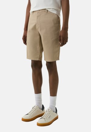 Shorts en coton beige à coupe droite, avec poches avant et fermeture à bouton. Associés à des chaussettes blanches à texture et à des sneakers blanches avec semelles en gomme.