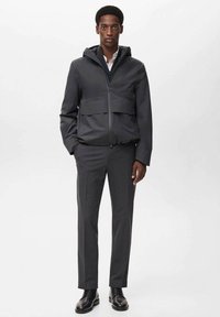 Jachetă gri închis cu glugă, dotată cu închidere cu fermoar și buzunare frontale cu clapetă, asociată cu pantaloni croiți asortați și pantofi negri.