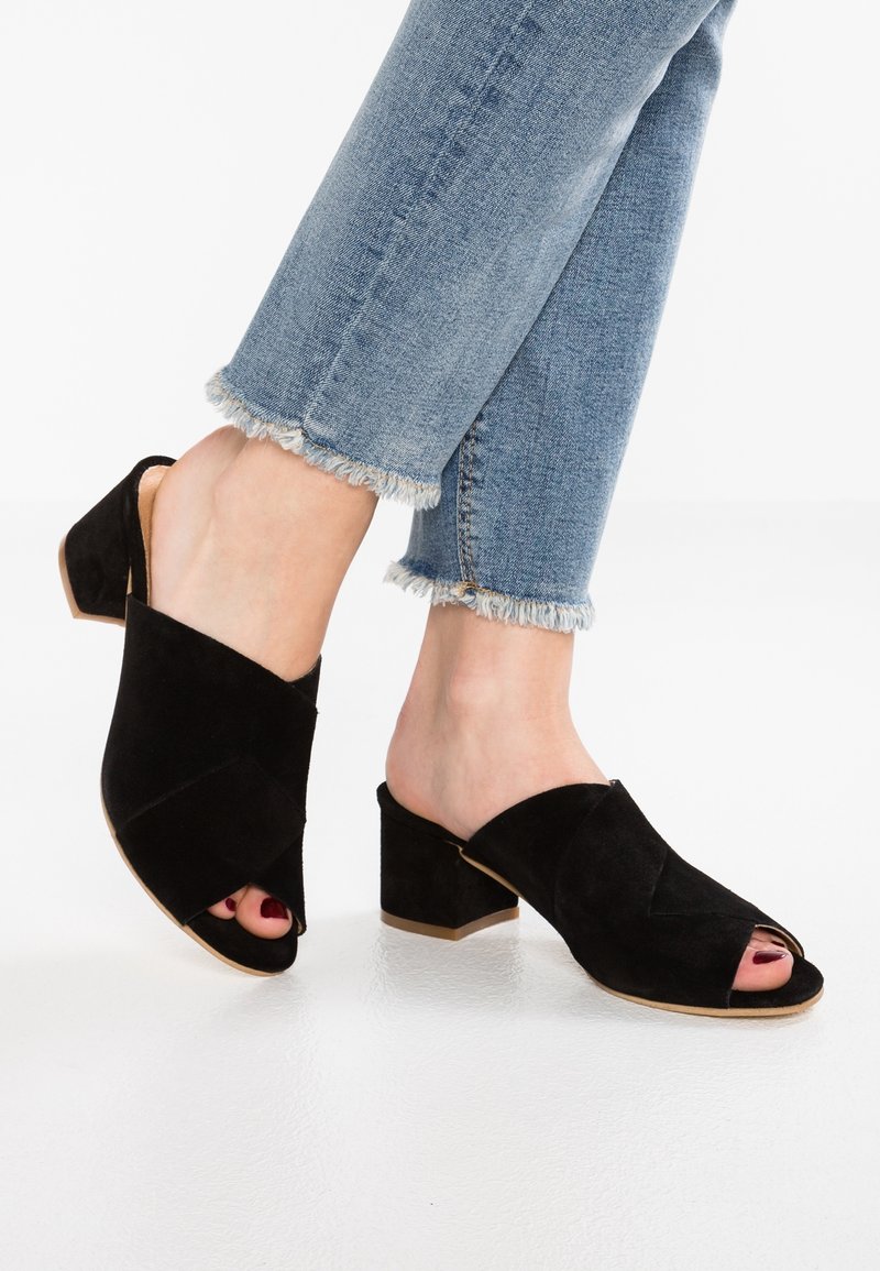 Mules en suède noir à bout ouvert avec un design entrecroisé et un talon bloc. Portés avec un jean à ourlet effiloché, mettant en valeur la texture et la forme de la chaussure.
