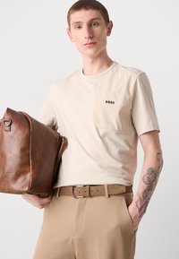 Beige rövid ujjú póló texturált zsebbel és "BOSS" logóval, világosbarna nadrággal és barna bőrtáskával párosítva.