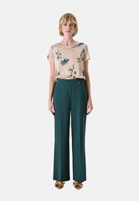 Roze bloemen blouse met korte mouwen, gecombineerd met hoge taille, wijdvallende groene broek. Gouden decoratieve details op de schoenen.