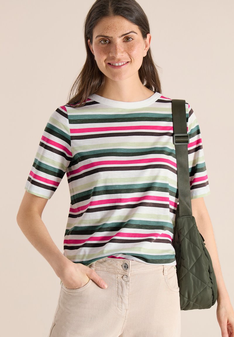 Femme portant une chemise à manches courtes rayée et un pantalon clair, portant un sac à bandoulière vert matelassé, souriant à la caméra.