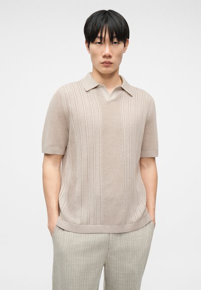 Νεαρός άνδρας με κοντά μαύρα μαλλιά, φορώντας μπεζ πλεκτό πόλο με κοντά μανίκια και ανοιχτό γκρι παντελόνι με λεπτές ρίγες, με τα χέρια στις τσέπες.